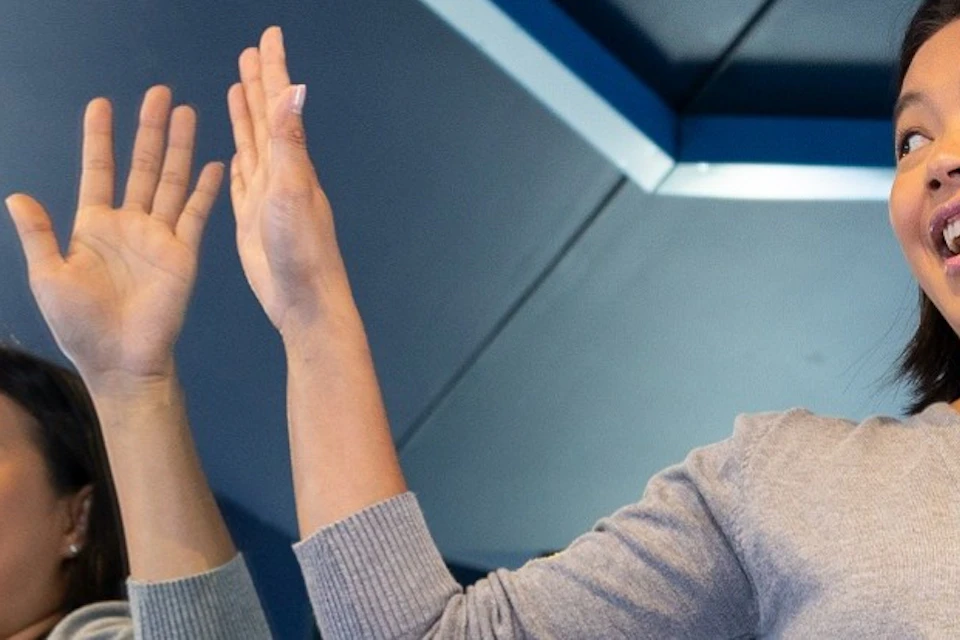 A woman sitting on a blue surface in a room with mirrors, creating multiple reflections. She raises her hands, smiling.