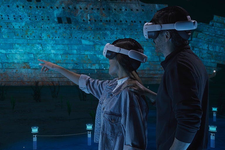 Dues persones amb auriculars de realitat virtual interactuant amb una escena submarina, amb un gran vaixell enfonsat al fons.