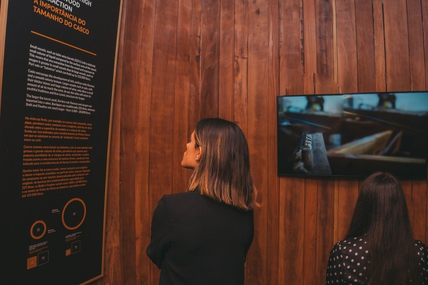 A person in a black shirt looks at a display board on a wooden wall, with a television screen showing an image to the right.