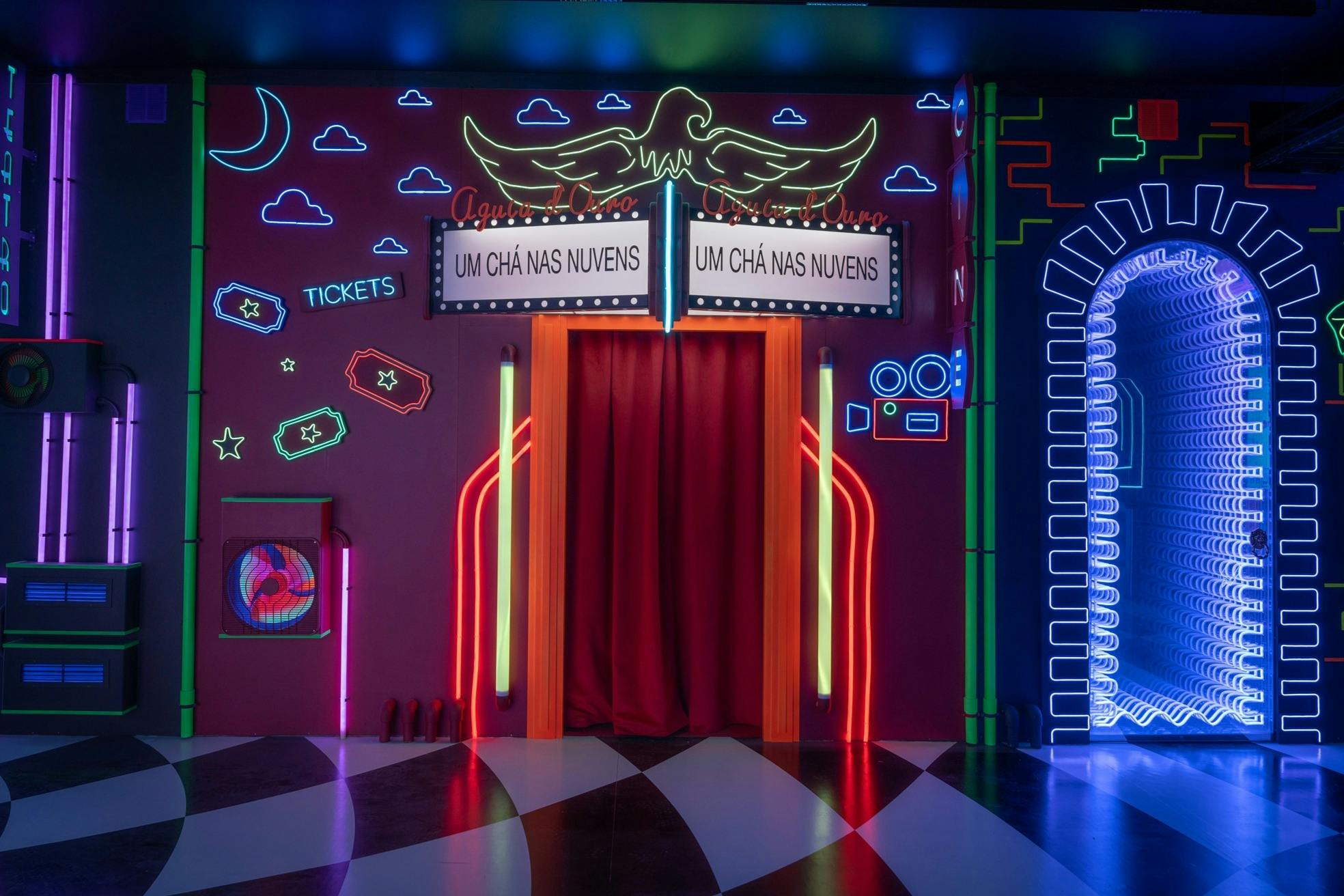 霓虹灯照明的入口配有红色窗帘，两侧是“Um Chá Nas Nuvens”标志，与彩色墙壁搭配各种霓虹灯设计。