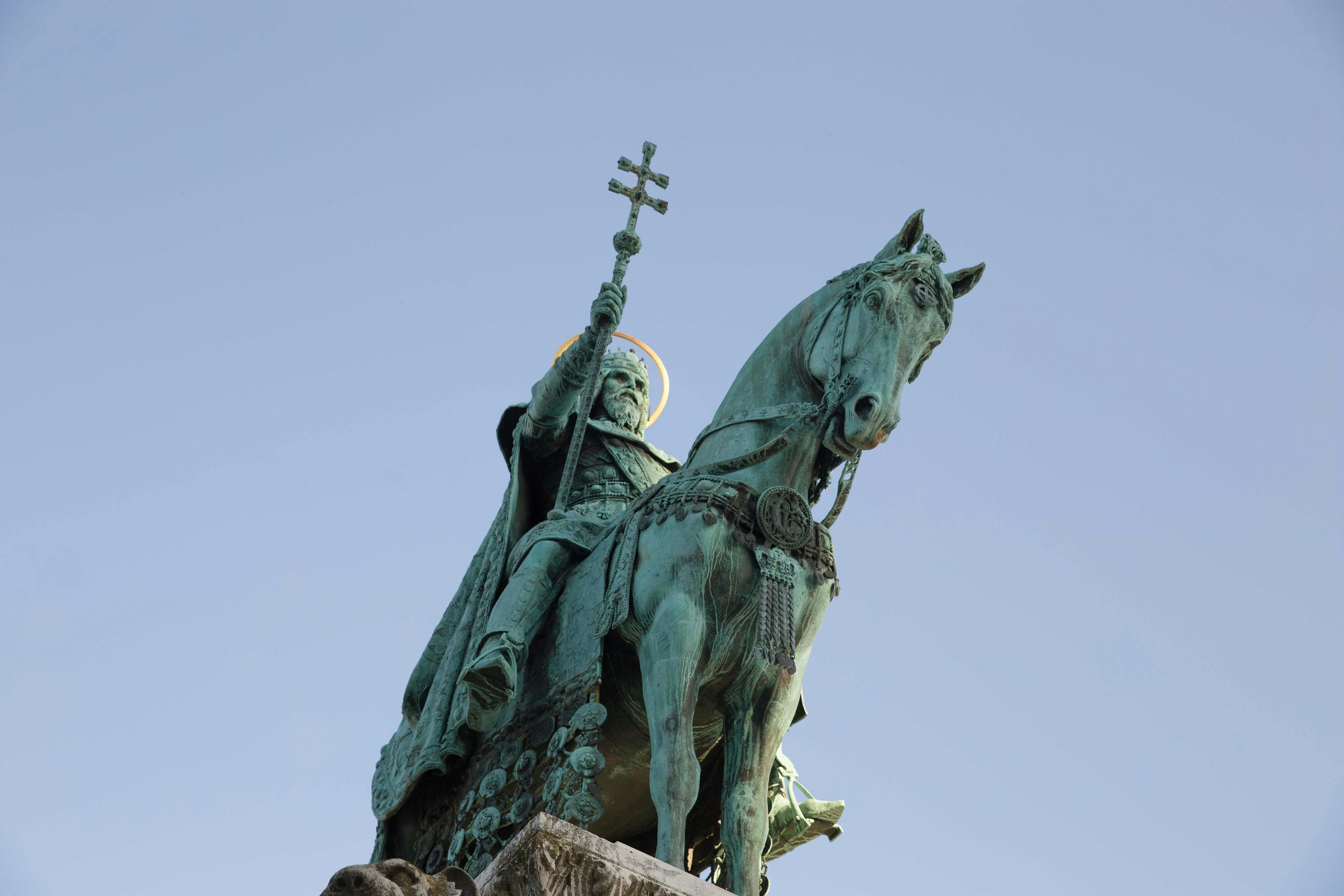 石の台座の上で、澄んだ青い空に向かって、十字架を持ったひげのある人物の青銅の馬術像。