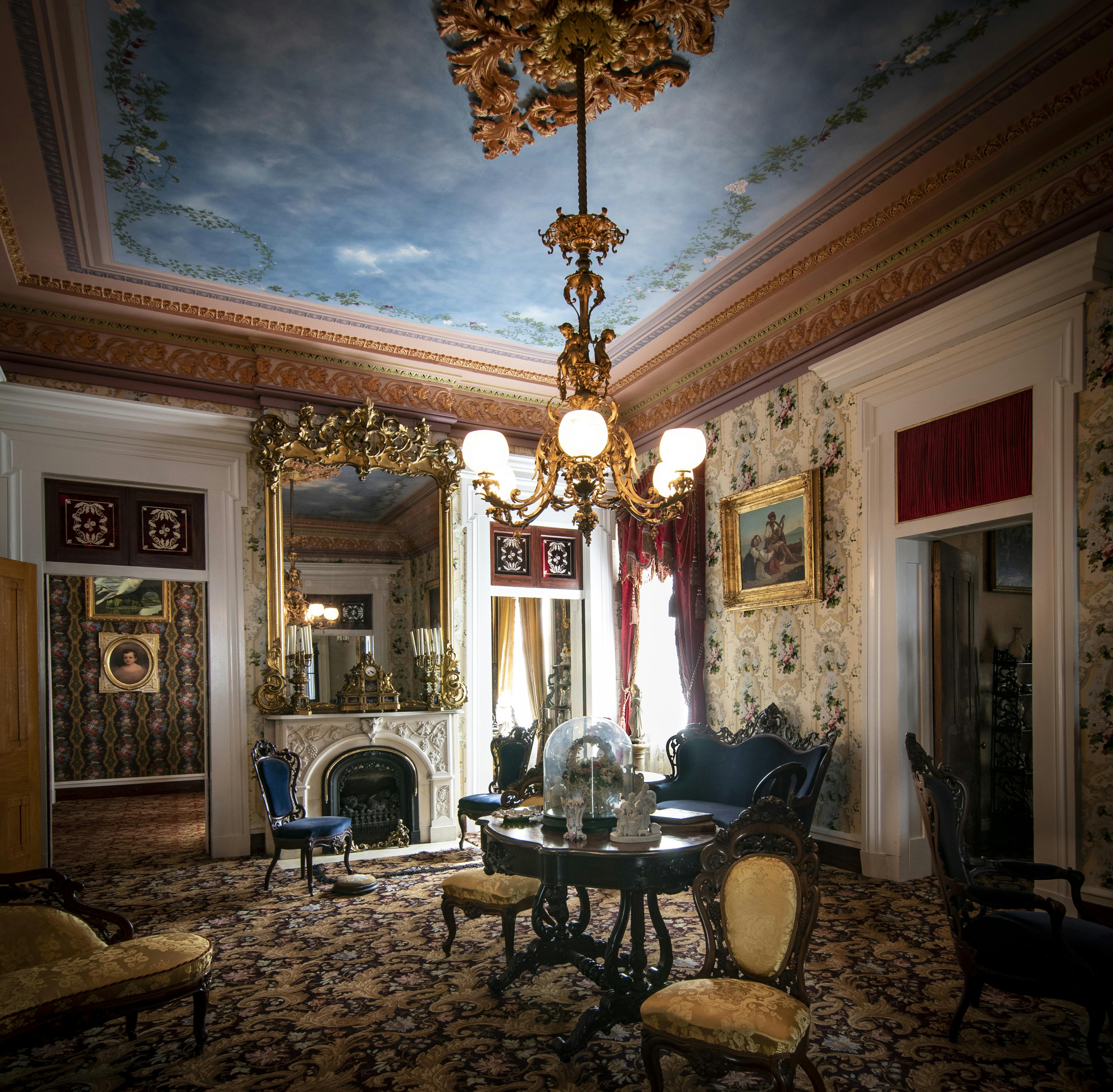 Κομψό δωμάτιο βικτοριανού στιλ με περίτεχνο χρυσό πολυέλαιο, μεγάλο καθρέφτη πάνω από το τζάκι, ταπετσαρία με σχέδια και έπιπλα αντίκες.