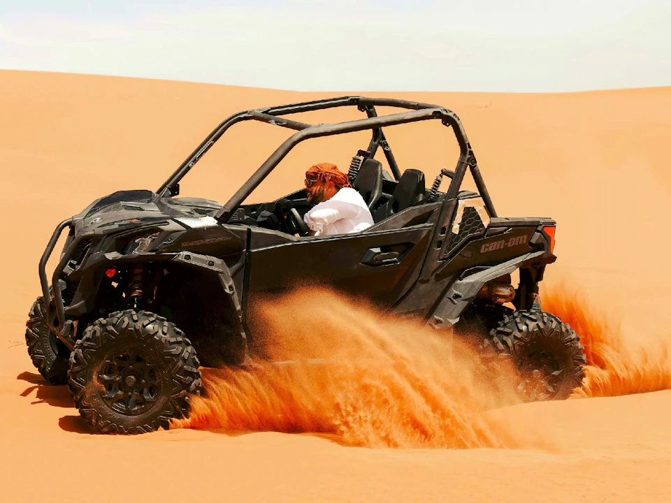 A person driving a black off-road vehicle through a sandy desert, with sand being kicked up by the tires.