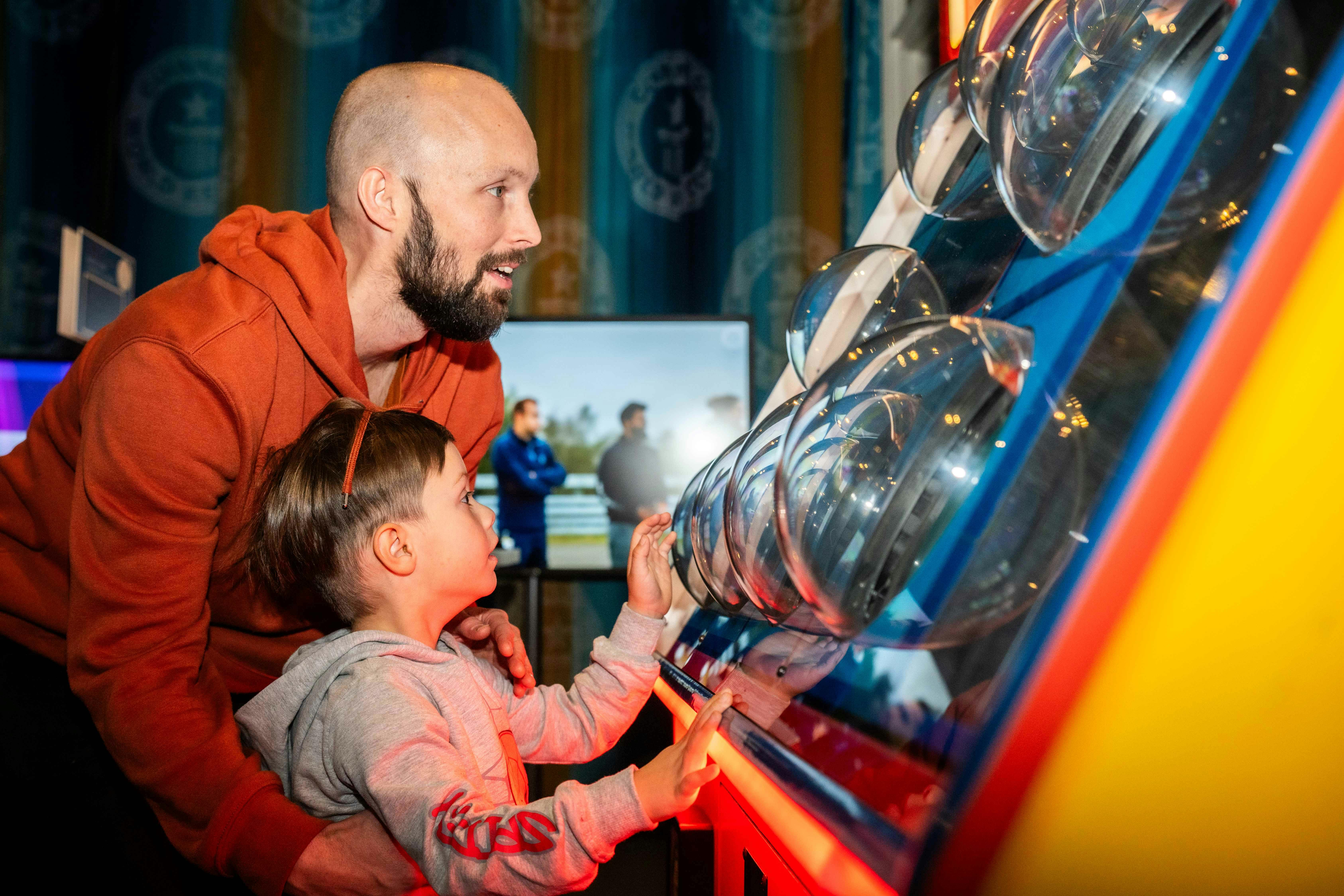 A man and a child, both focused, interact with a brightly lit, circular exhibit game indoors.