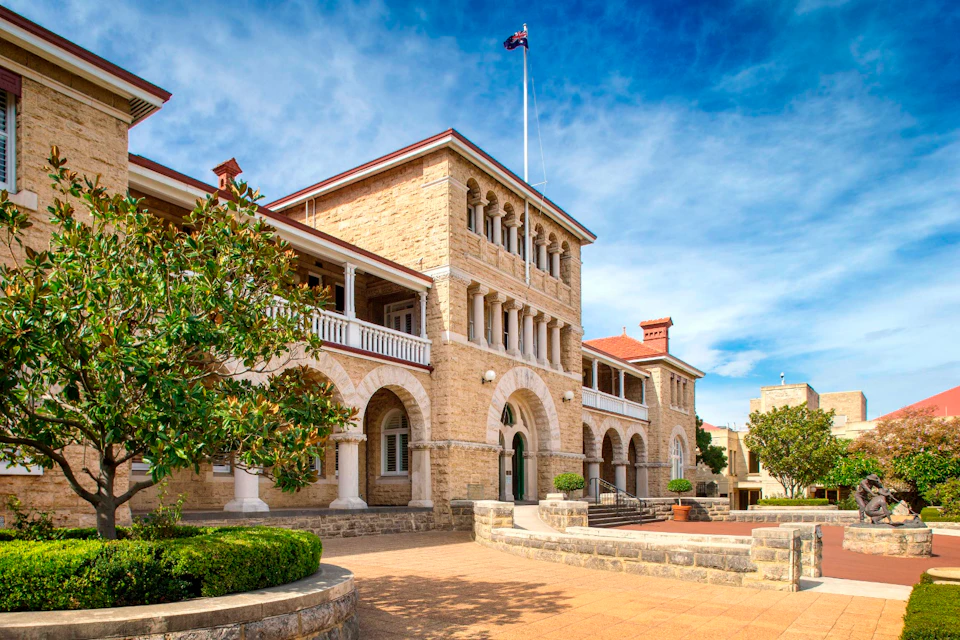 A historic stone building with arches and a flagpole, set against a blue sky with trees and a courtyard in the foreground.