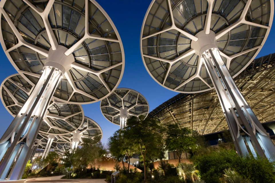 Large, futuristic, umbrella-like structures with lights and greenery underneath set against a twilight sky.