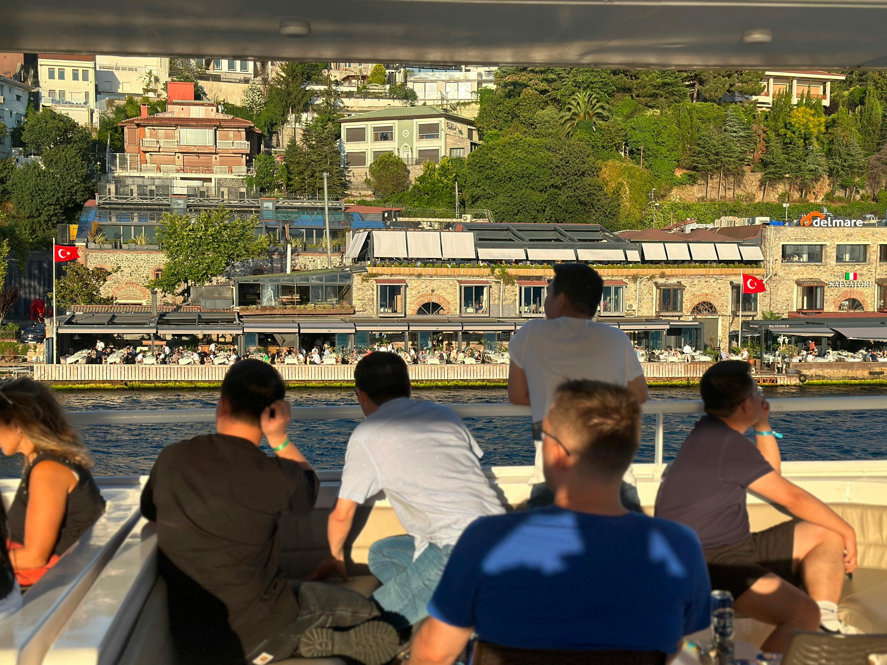 Five people sitting on a boat look towards a shoreline with buildings, trees, and a Turkish flag visible.