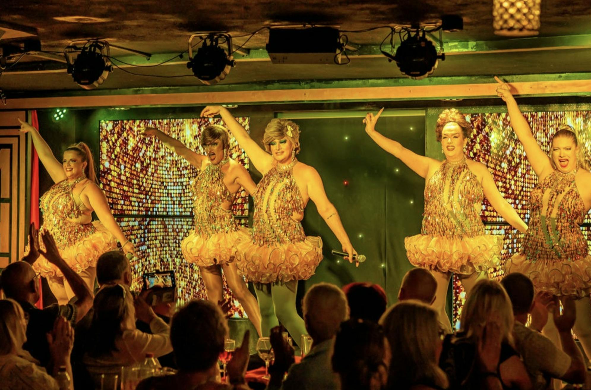 Исполнители в сверкающих костюмах танцуют на сцене при ярком освещении, на переднем плане сидит зритель.