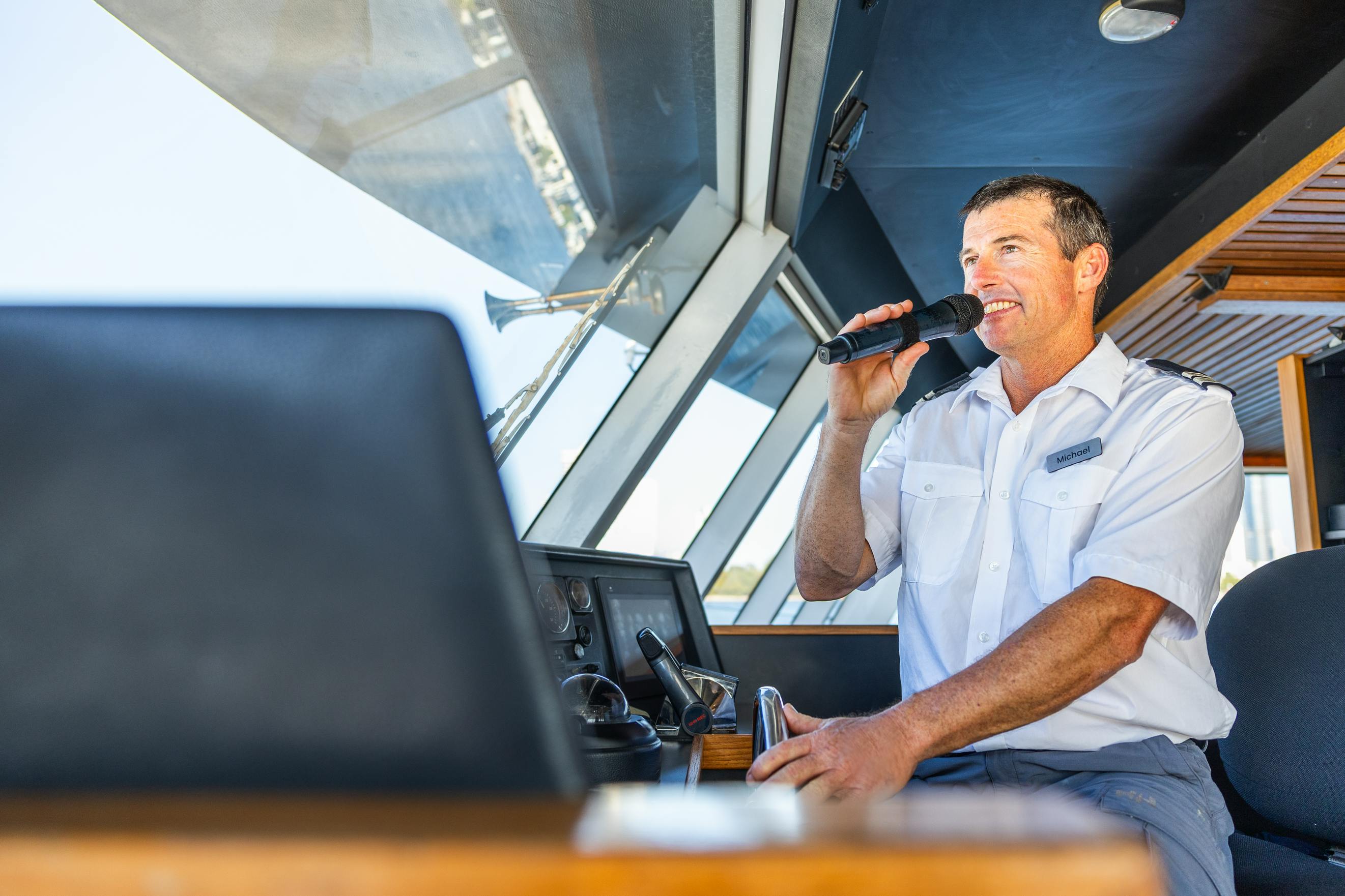 Ένας ένστολος μιλάει σε ένα μικρόφωνο στο τιμόνι μιας βάρκας με μεγάλα παράθυρα πίσω του.