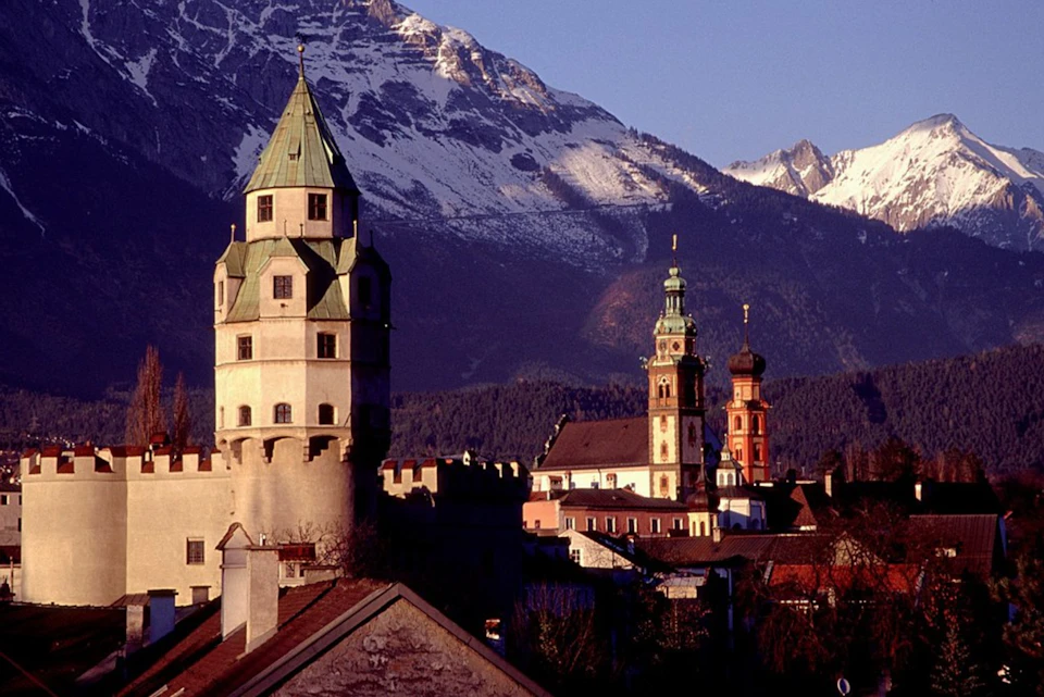 Snow-capped mountains with a medieval tower, church spires, and rooftops of a historic town in the foreground.