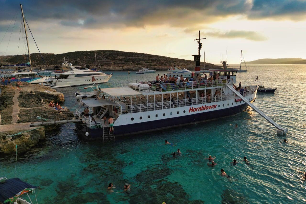 Ein Boot mit dem Namen "Hornblower" hat im klaren türkisfarbenen Wasser angelegt, mit schwimmenden Menschen in der Nähe und Hügeln im Hintergrund.