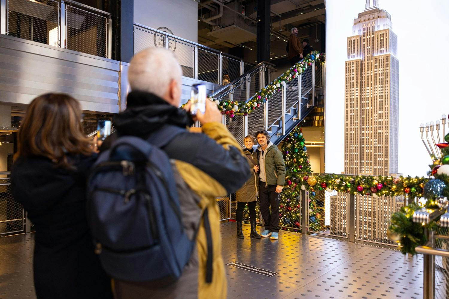 A person photographs a couple posing by Christmas decorations in an indoor setting with a model of the Empire State Building.