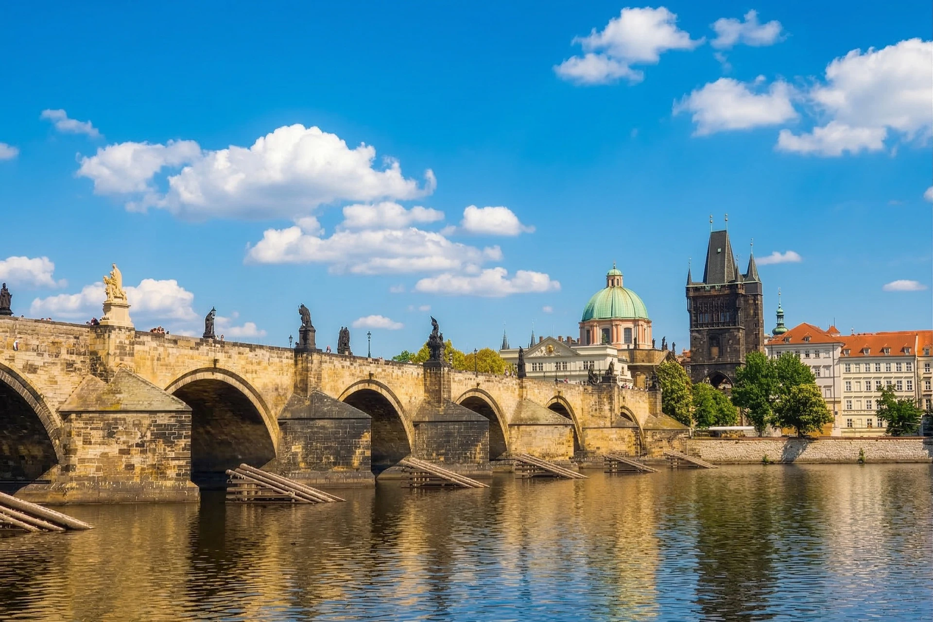 A historic stone bridge with statues spans over a calm river, with domed and spired buildings under a blue sky with clouds.
