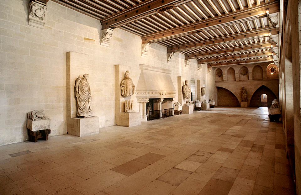 Spacious hall with stone statues, wooden beamed ceiling, large fireplace, and arched alcove in the background.