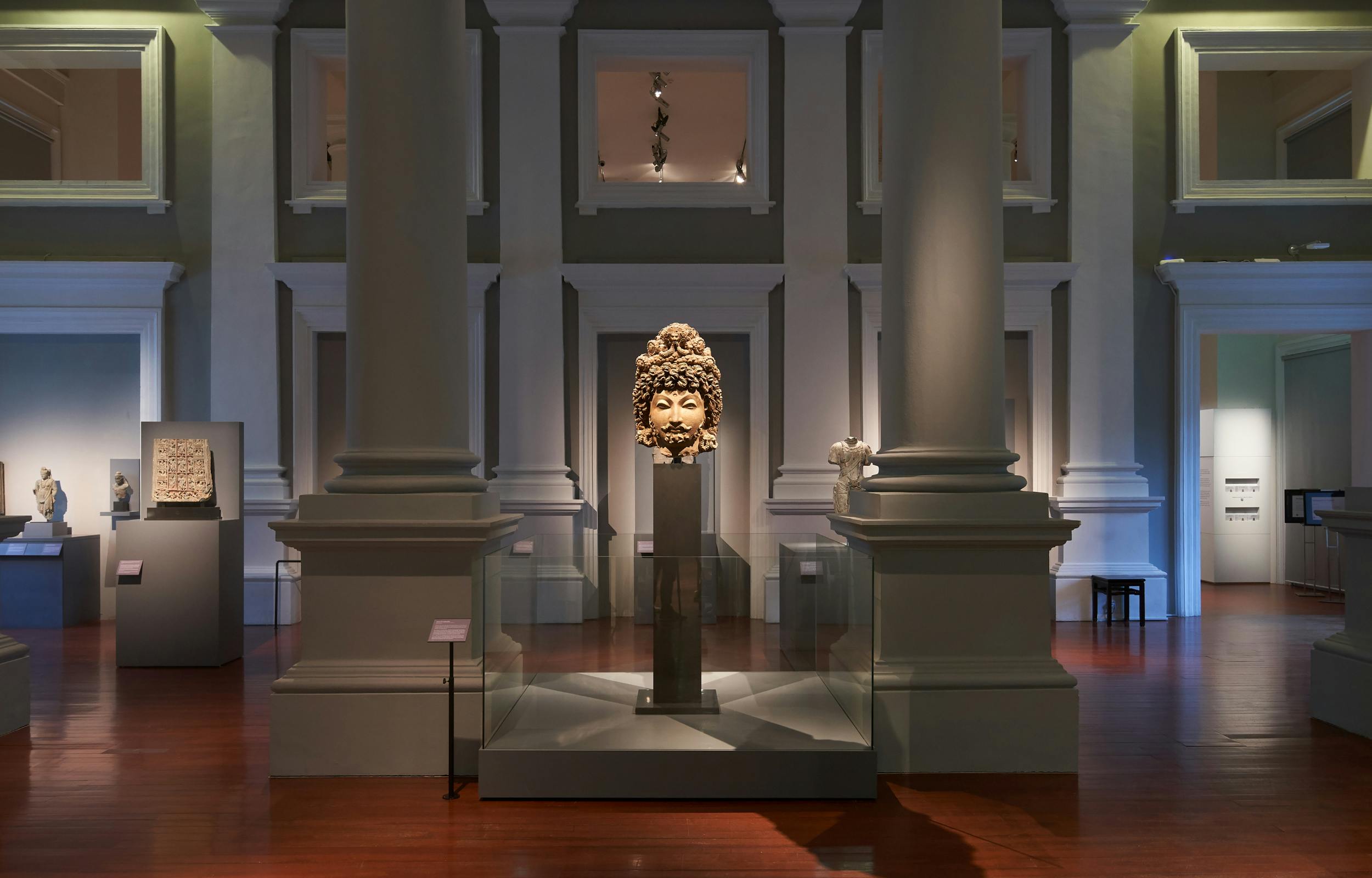 A museum exhibit featuring a sculpted head on a pedestal, enclosed in glass, surrounded by classical architecture.