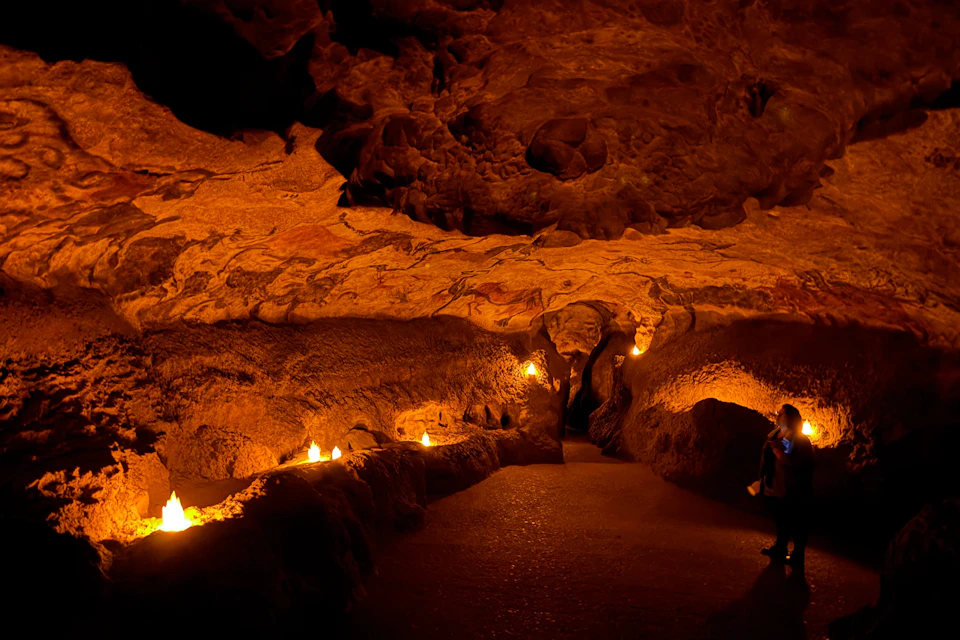 A dimly lit cave interior with a winding path and illuminated by small lights; a person stands on the right side observing.