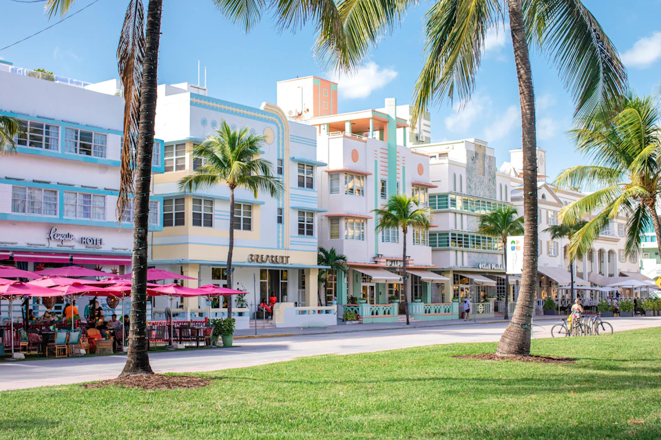 A row of pastel-colored Art Deco buildings with palm trees in front, seen from a grassy area under a sunny sky.