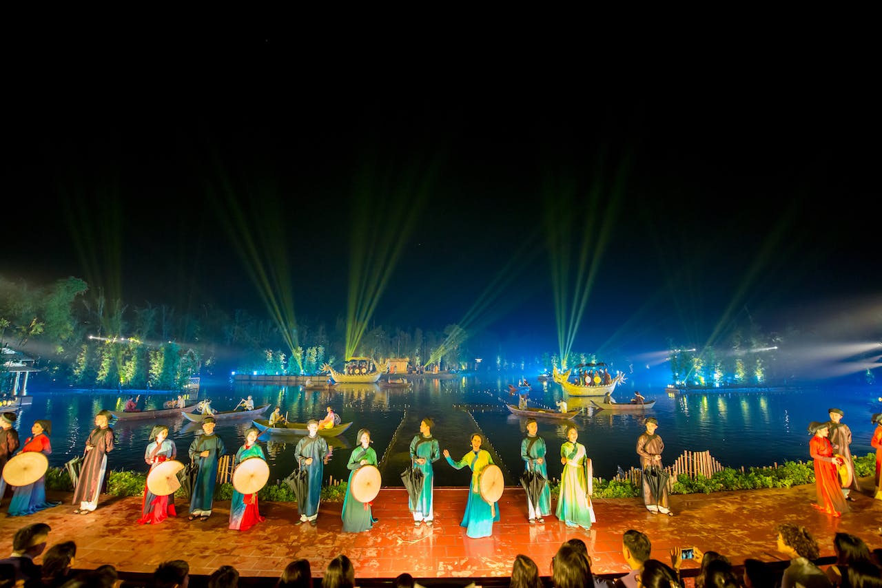 Darsteller in traditioneller Tracht auf der Bühne, im Hintergrund bei Nacht beleuchtete Boote und Lichtstrahlen über dem Wasser.