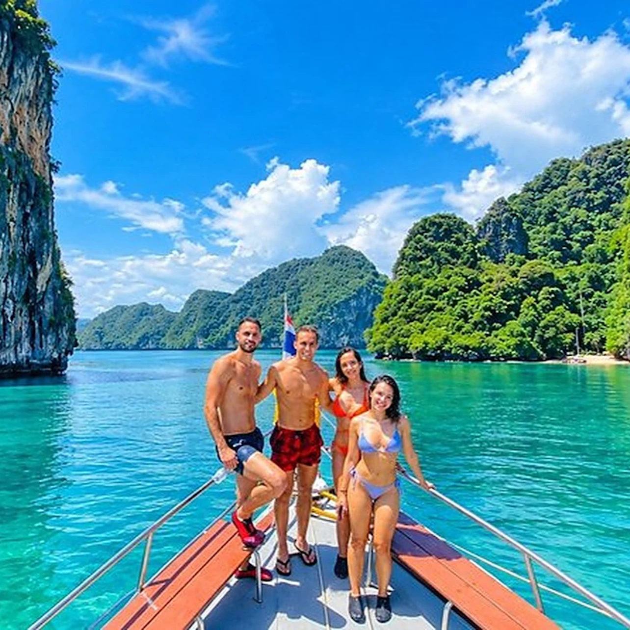 Four people in swimwear stand on a boat with vibrant green water, limestone cliffs, and a sunny blue sky in the background.