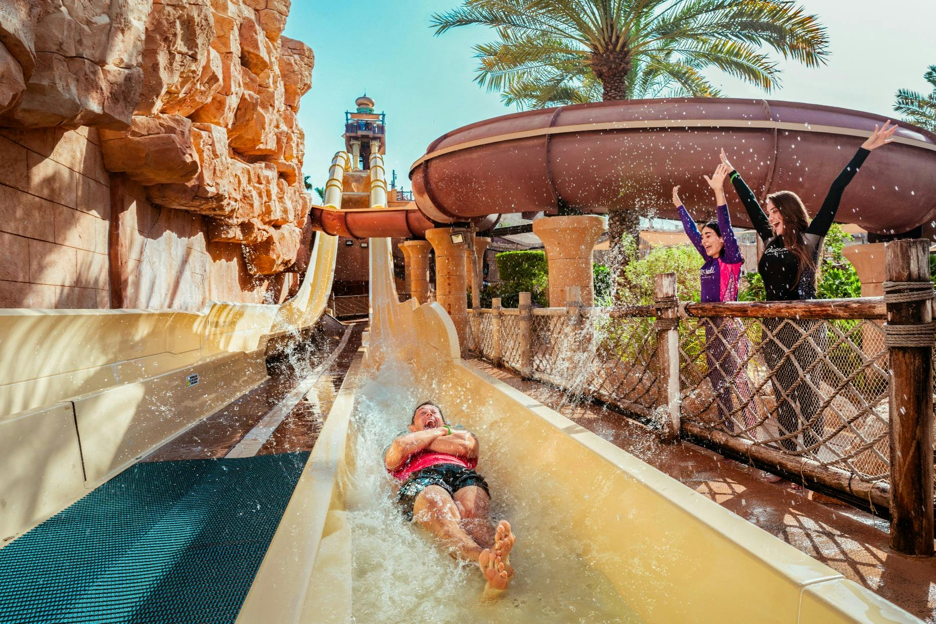 Une personne glissant sur un toboggan aquatique dans un parc aquatique, entourée de spectateurs et de palmiers.