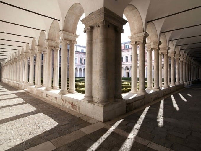 Каменный дворик с аркой колонн, отбрасывающей тени на каменно-плиточный грунт. Ухоженные кусты и здание на заднем плане.