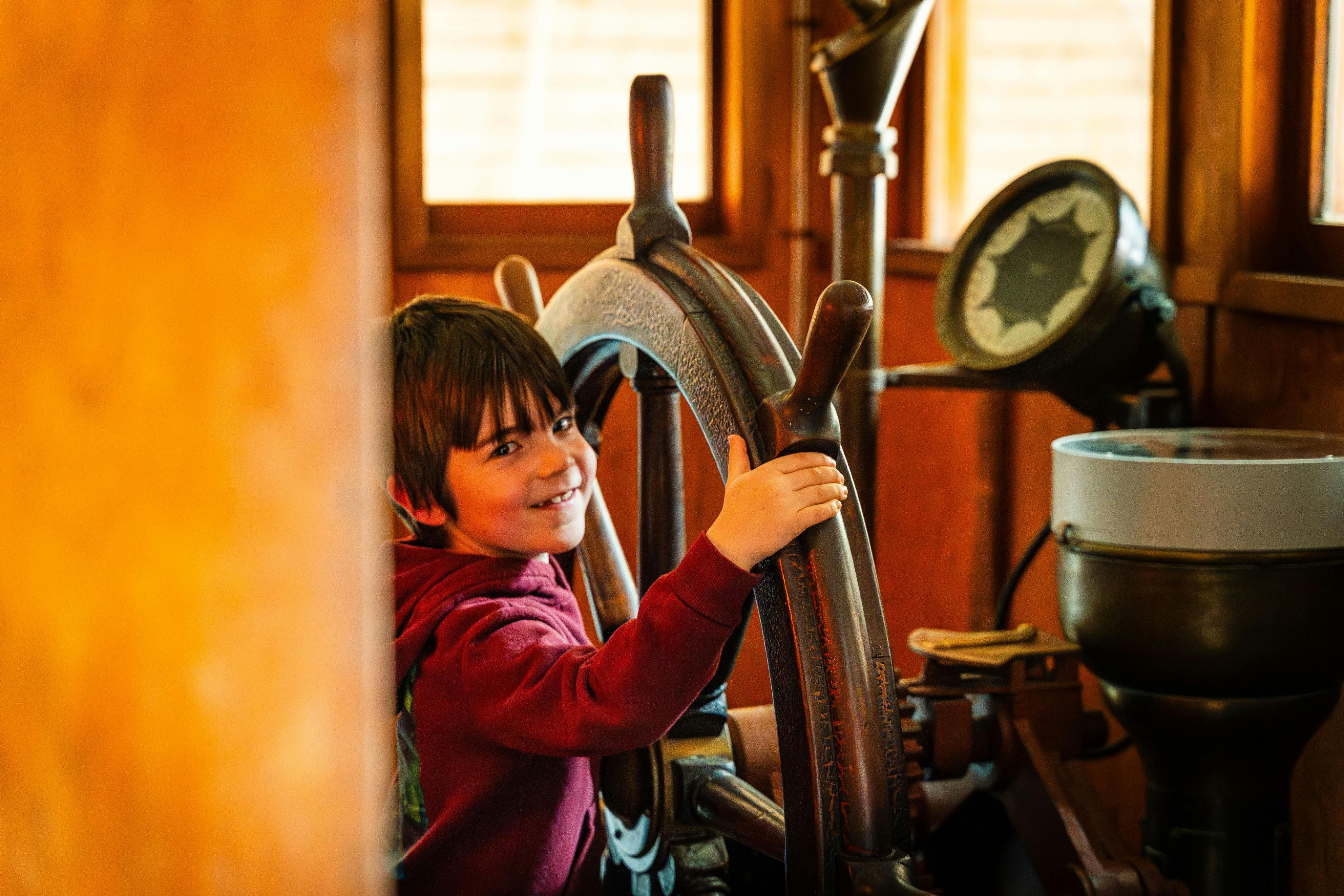 Et barn i rød hættetrøje smiler, mens det holder et stort skibshjul i træ i et varmt oplyst interiør.