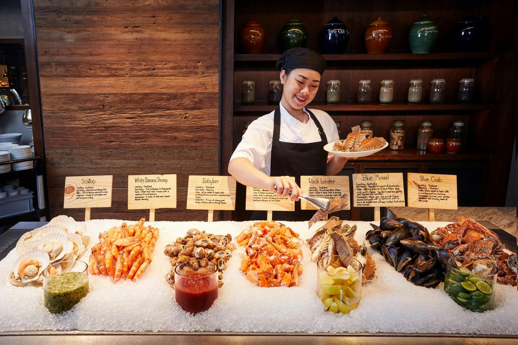 A chef arranging seafood, including shrimp, lobster, and mussels, on ice at a buffet with labeled signs and jars in the background.