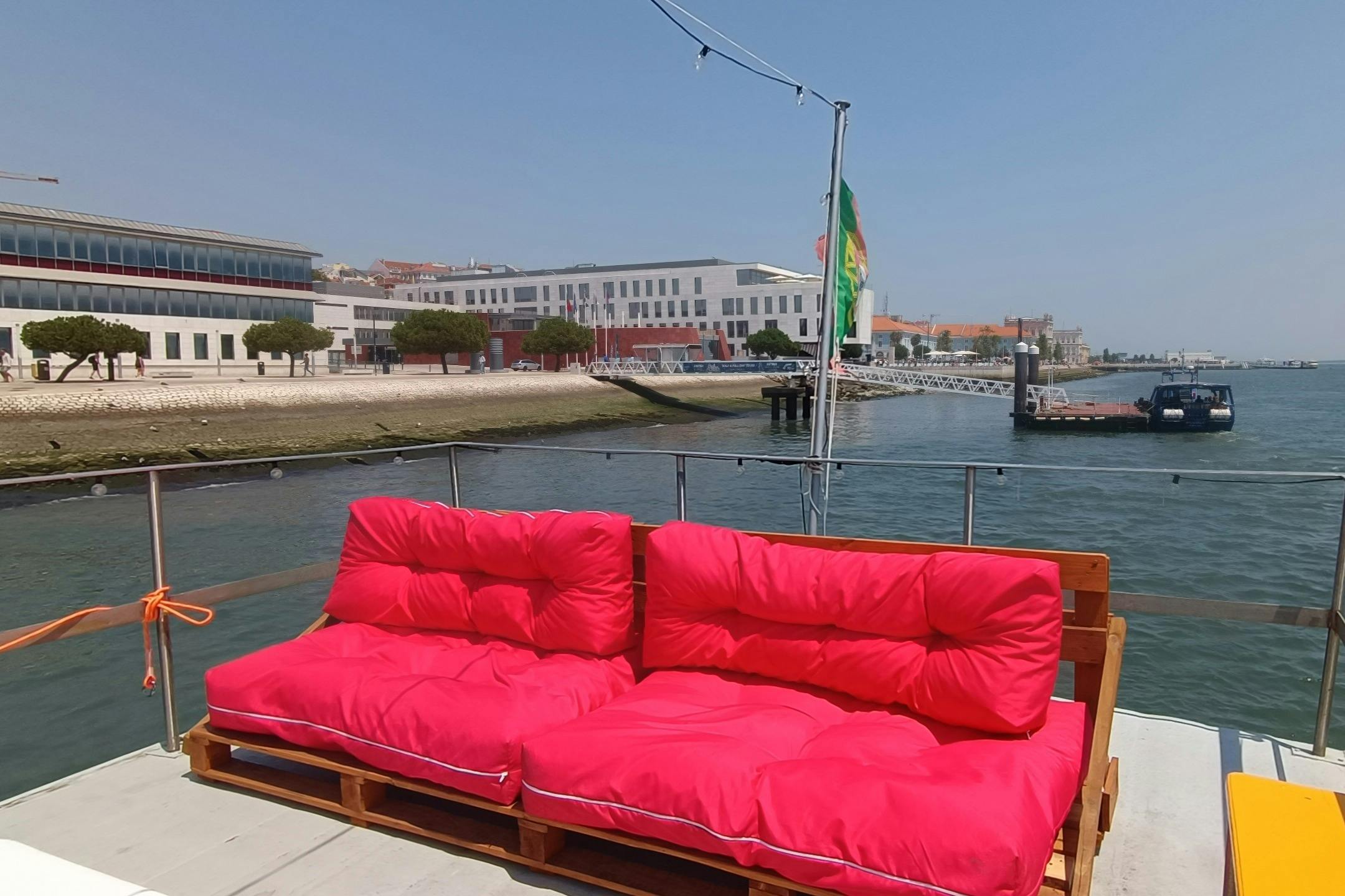 2つの明るいピンクのクッション付きパレットは、風光明媚なウォーターフロントと建物を背景にしたドッキングボートの座席として機能します。