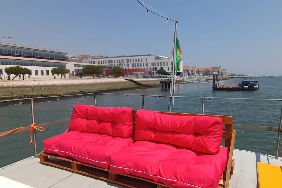 Two bright pink cushioned pallets serve as seating on a docked boat with a scenic waterfront and buildings in the background.