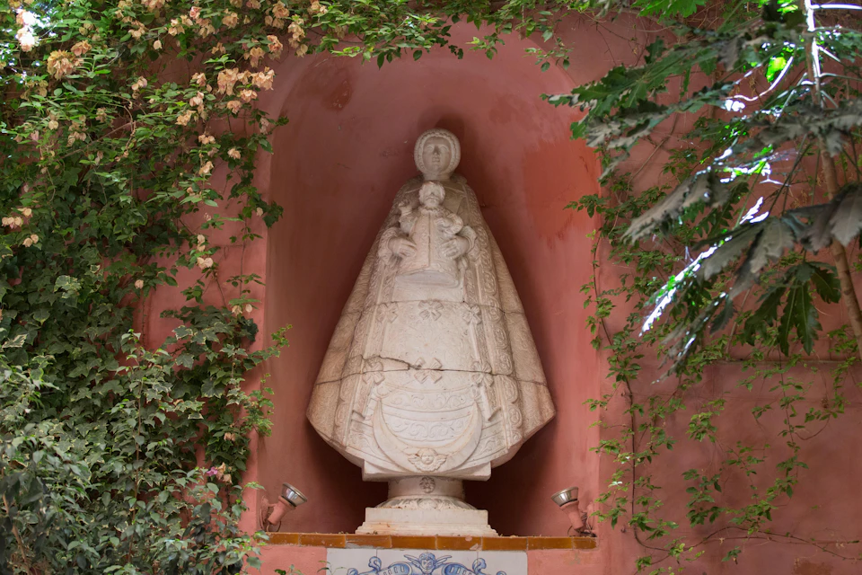 A stone statue of a robed figure holding a child is set in a niche surrounded by plants and flowers.