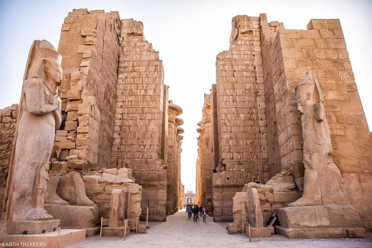Ancient stone ruins with towering walls and large statues. People are walking down the central path between the structures.