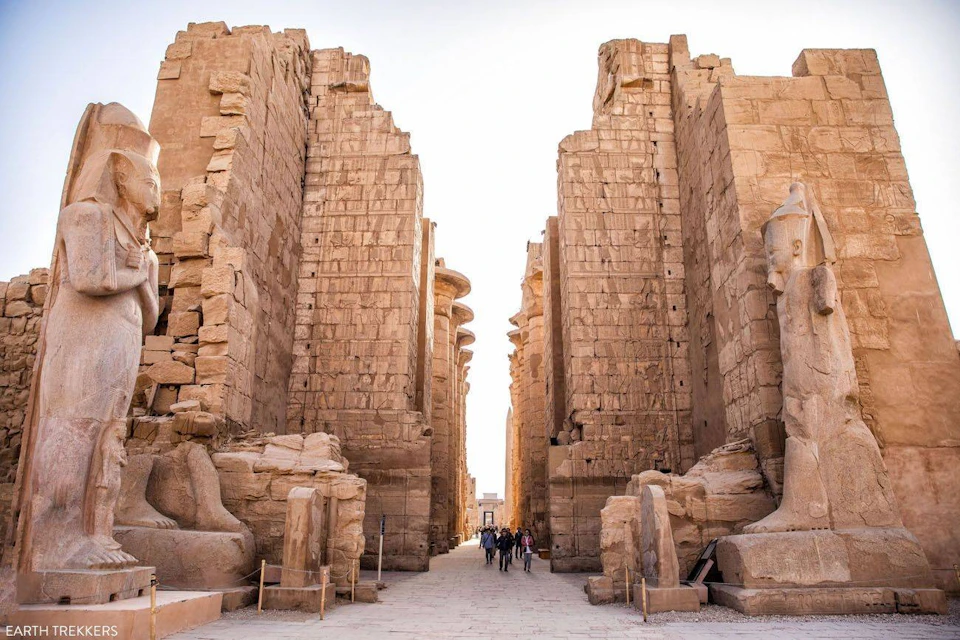 Ancient stone ruins with towering walls and large statues. People are walking down the central path between the structures.
