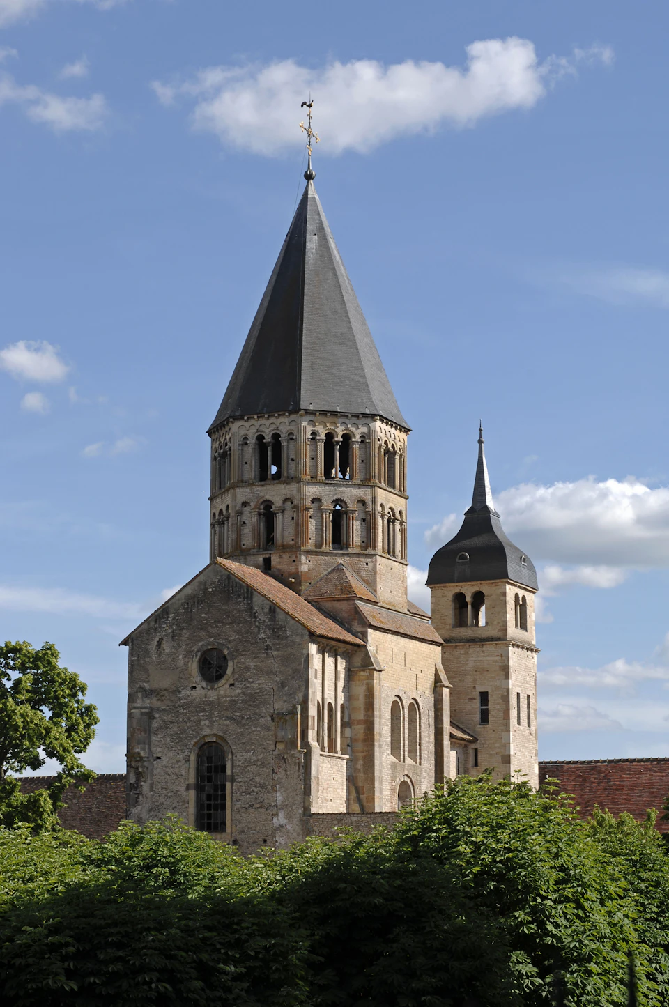 Stone church with a tall, pointed central tower and a smaller round bell tower against a blue sky with a few clouds.