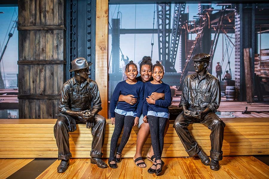 Tre bambini seduti tra due statue di operai su una panchina di legno, con impalcature industriali sullo sfondo.