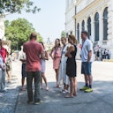 A group of people standing and listening to a guide outside a large, ornate building on a sunny day. Trees and a statue are visible in the background.