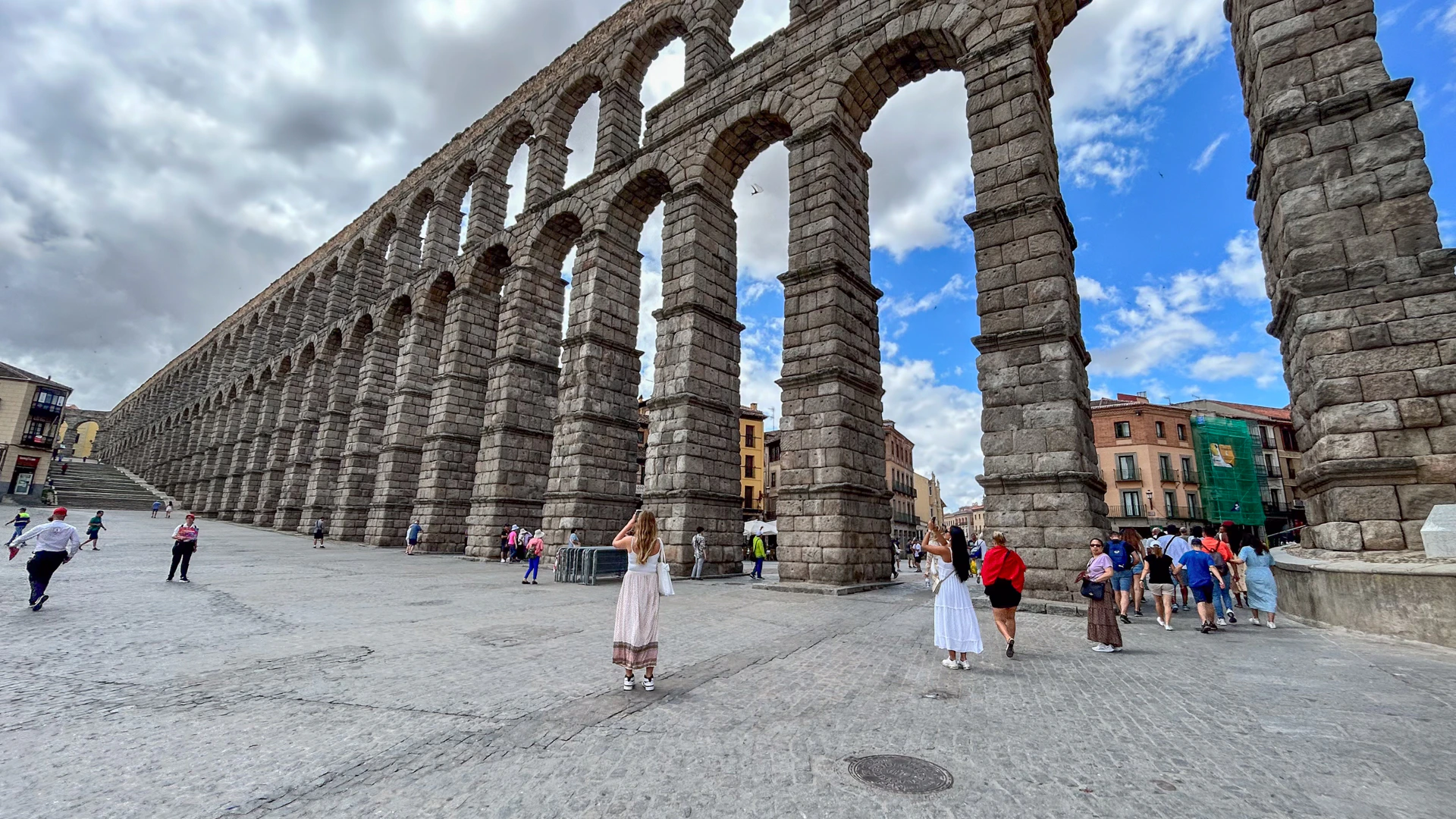 People walking near an ancient stone aqueduct with arches, under a partly cloudy sky in a city plaza.