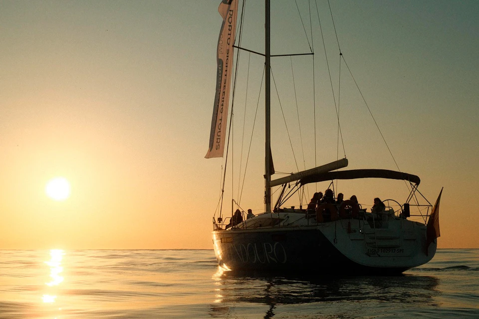 A sailboat with a group of people on board floats on calm water at sunset.