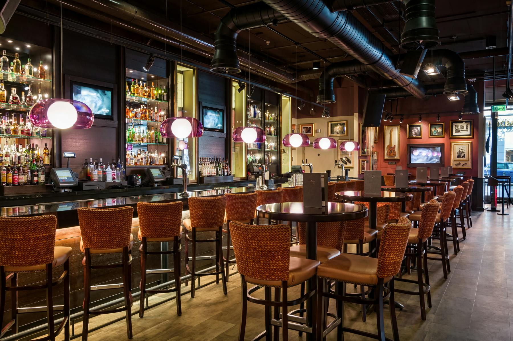 A stylish bar with tall wicker chairs, round tables, hanging spherical lamps, and an assortment of bottles behind the counter.