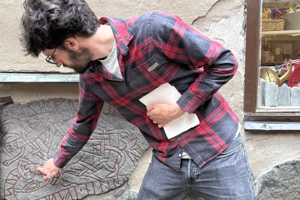 A man in a red plaid shirt points at a stone carving with intricate patterns, holding a white sheet of paper in his other hand.