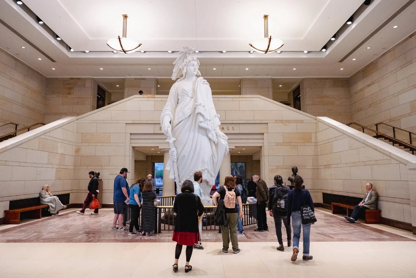 在一个宽敞、光线充足、大理石墙壁和高高的天花板的大厅里，一群人欣赏着一尊白色长袍雕像。