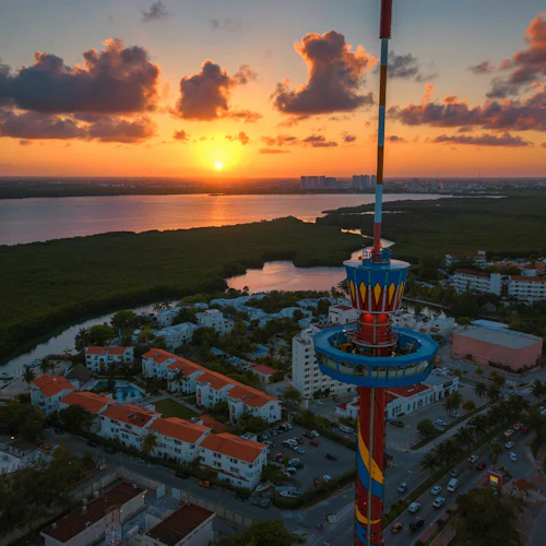 Ein hoher Aussichtsturm mit roten und gelben Farben steht bei Sonnenuntergang über einem Viertel mit weißen Gebäuden und roten Dächern.