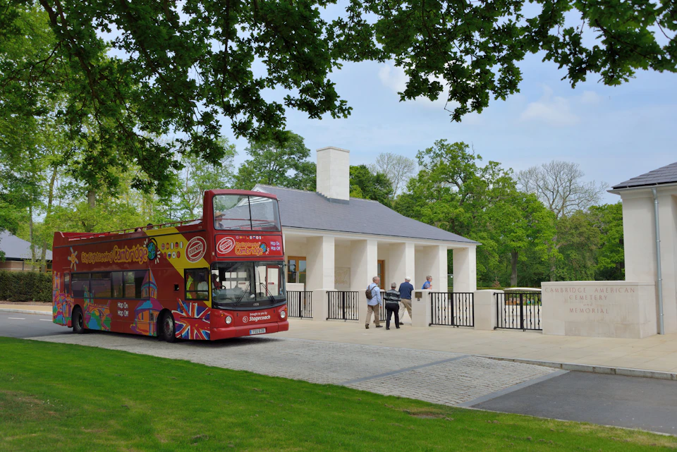 A red open-top tour bus is parked near a white building; a few people stand nearby under leafy trees.