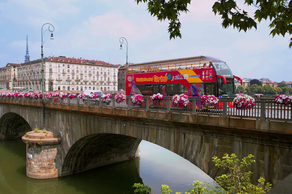 A red double-decker bus crosses a flower-adorned bridge over a river, with buildings and streetlights in the background.
