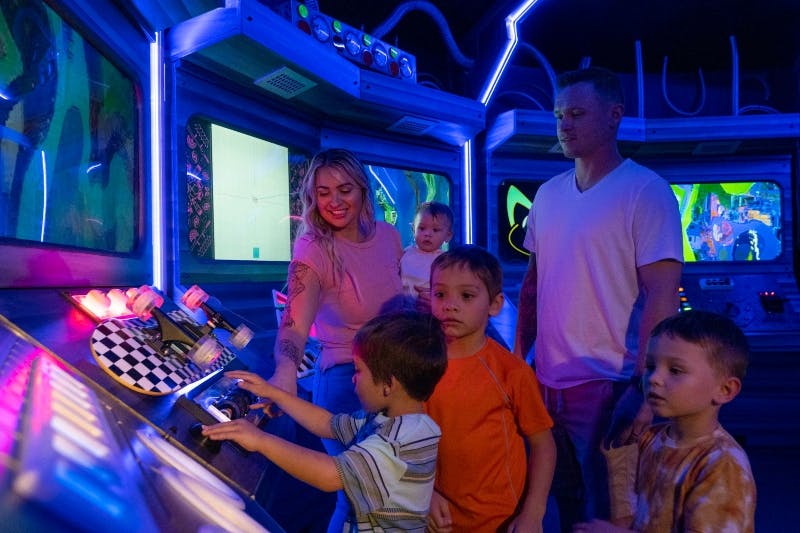 A family with young children interacts with colorful, futuristic controls and screens in a themed, illuminated environment.