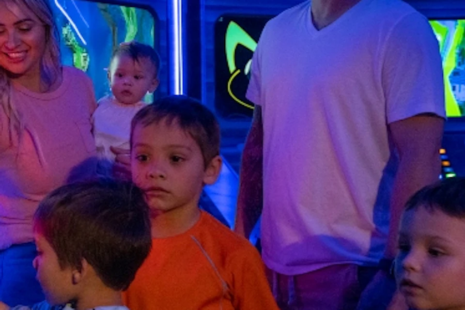 A family with young children interacts with colorful, futuristic controls and screens in a themed, illuminated environment.