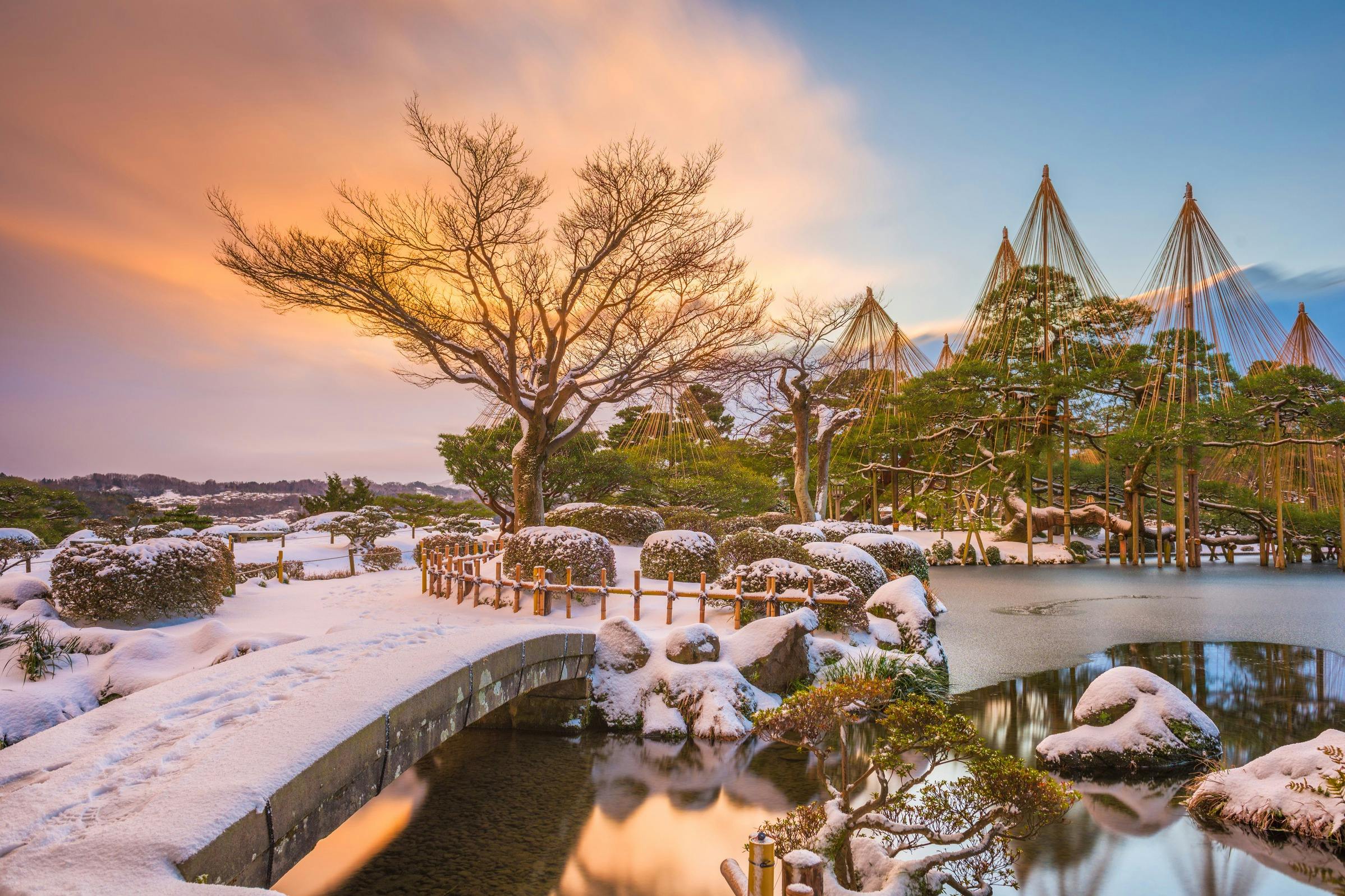 A snow-covered traditional Japanese garden at sunset, with trees, a stone bridge, and a pond reflecting the warm light.