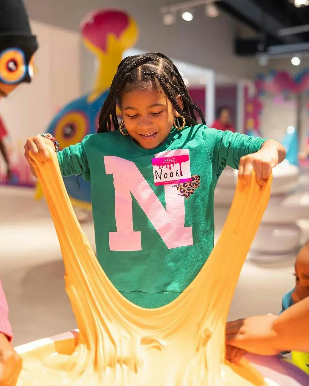 Dítě v zelené košili s velkým „N“ a jmenovkou, Noor, se usmívá, zatímco drží pružnou žlutou látku.