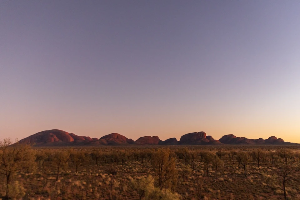 A wide view of a desert landscape with distant rocky hills at sunset, under a clear sky with a single star.