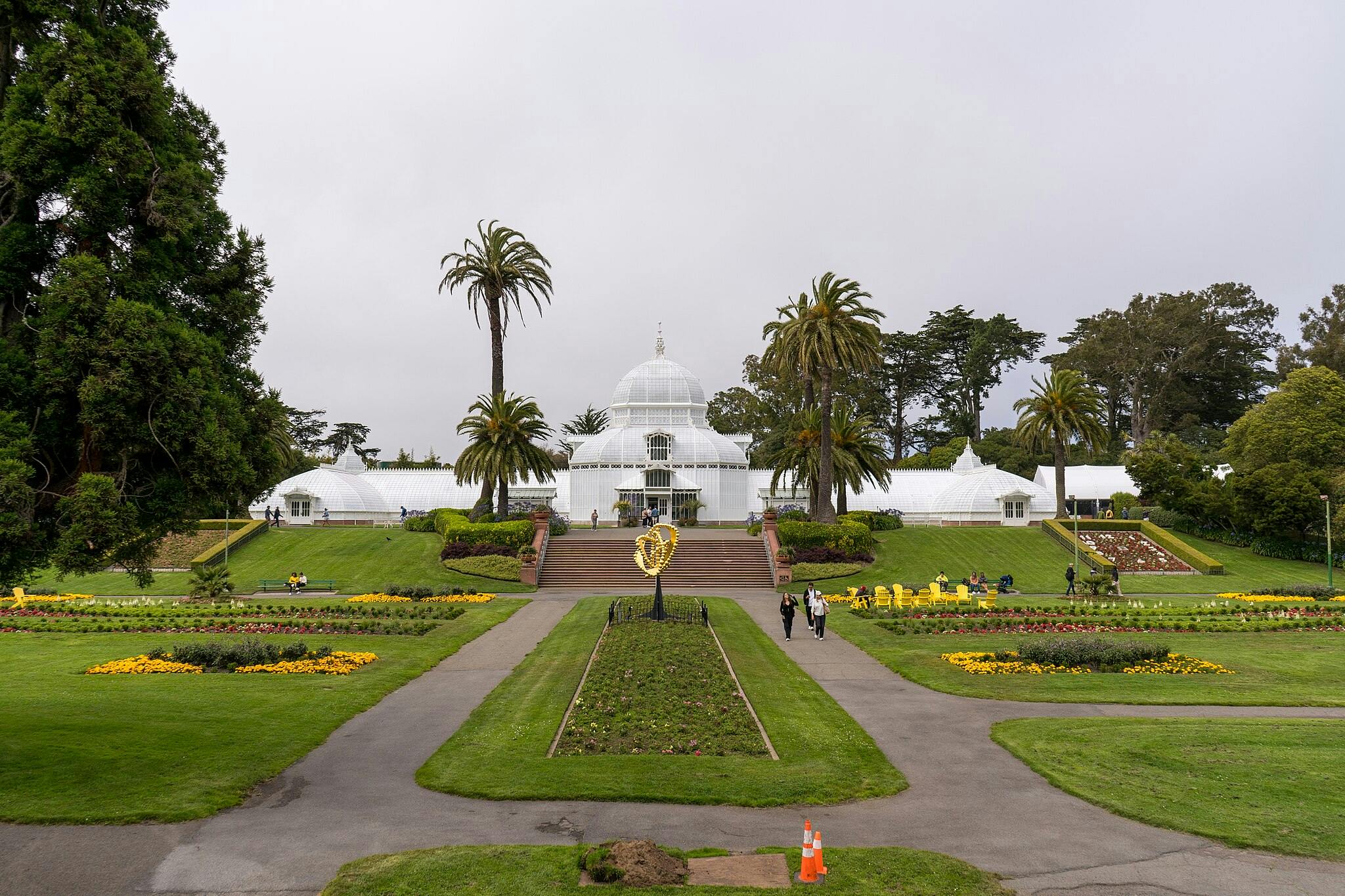 Symetrická zahrada s květinovými záhony a palmami vedoucí ke skleníku s bílou klenbou. Lidé chodí a sedí na lavičkách.