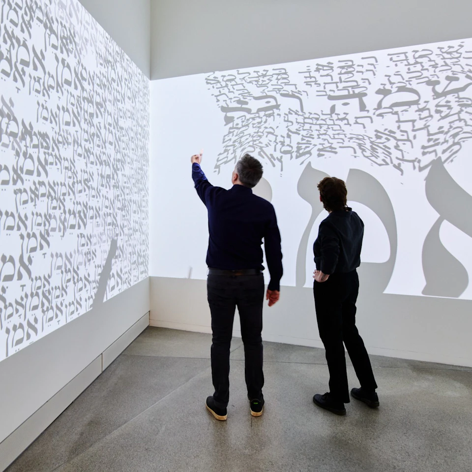 Two people stand in a room examining projections of calligraphy on white walls. One person points toward the text.